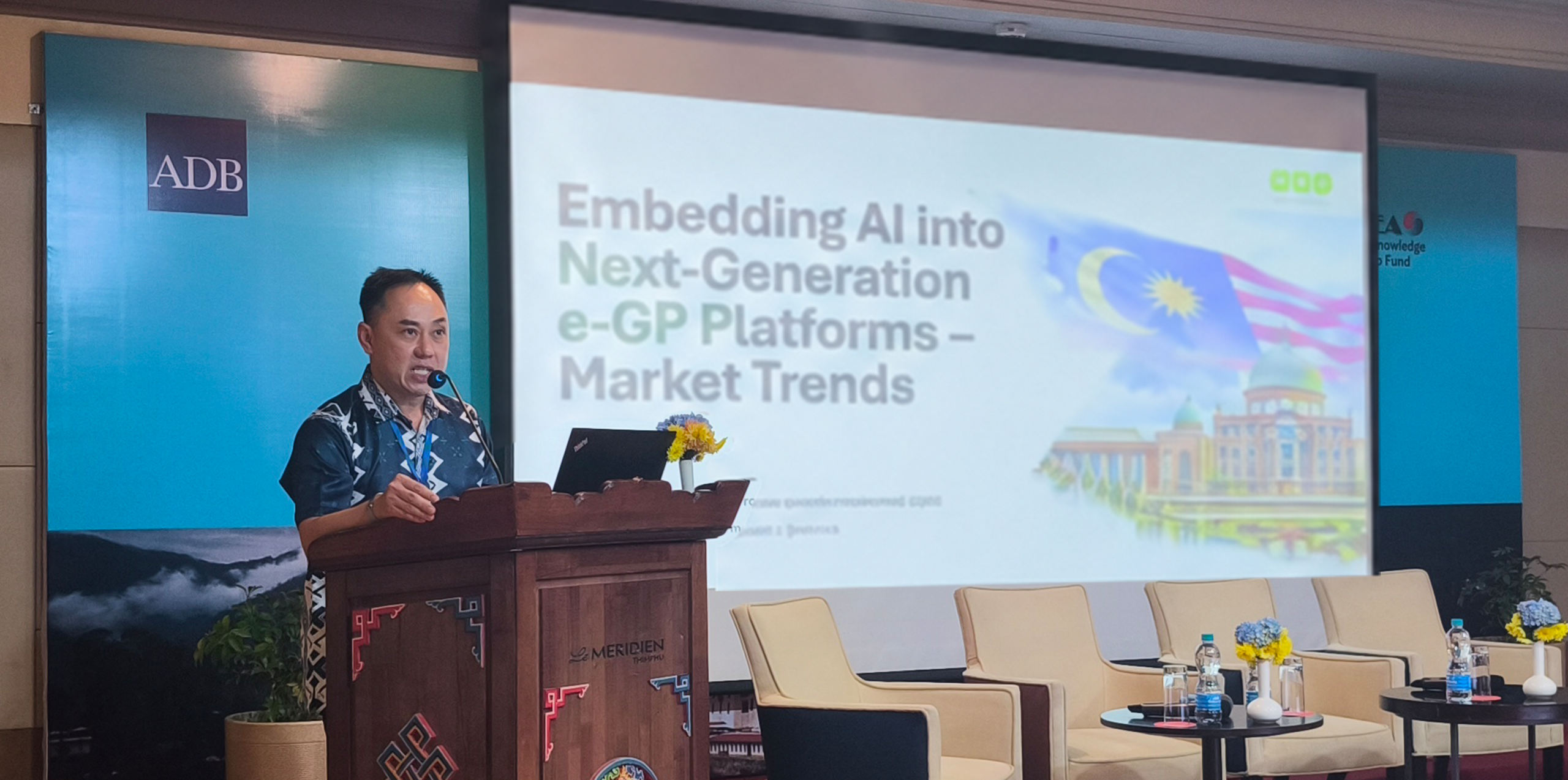 Melvin Loh presenting at a lectern beside a screen displaying AI and electronic government procurement content at APPeN 2026.